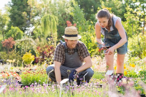 A woman and a young girl are working in a well-maintained garden, with the woman kneeling on the grass and the girl leaning forward, both wearing casual clothes and garden gloves. The garden features colourful flower beds with pink, purple, and yellow blooms, bordered by low shrubs and a neatly trimmed hedge in the background. Behind them, tall trees with lush green foliage provide shade and natural scenery. The lawn area is vibrant and healthy, with a mix of dense grass and flower patches, while the soil beds are tidy and well-kept. Sunlight filters through the trees, casting a bright, natural light on the scene, suggesting a clear day suitable for outdoor gardening activities. This outdoor space shows careful planning and maintenance typical of professional gardening services, such as those offered by Landscapers Chiswick, supporting sustainable and eco-friendly gardening practices in the local area near Chiswick or West London.
