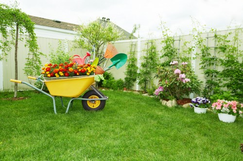 A well-maintained residential garden featuring a lush green lawn with uniform grass, bordered by a white fence covered in climbing plants and greenery, creating a natural enclosure. In the foreground, a yellow wheelbarrow is filled with colourful garden flowers, including red, yellow, and orange blooms. Nearby, a collection of garden tools such as a watering can, garden rake, and small spade are arranged, indicating ongoing gardening activity. Along the right side, there are flower beds with blooming pink, purple, and white flowers, bordered by soil and mulch, adding vibrant colour to the outdoor space. In the background, small trees and shrubs provide height and structure to the garden, while the overall scene is illuminated by natural daylight under a partly cloudy sky, reflecting a typical outdoor gardening environment. The layout suggests an organized landscaped yard designed for gardening and outdoor leisure, consistent with services offered by landscapers in Chiswick.