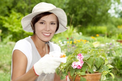 The image depicts a smiling woman in a white hat, gardening outdoors in a well-maintained garden. She is wearing a white top and gardening gloves, and she appears to be tending to a potted plant with pink flowers and green foliage. The background shows a lush, green environment with various plants, shrubs, and trees, suggesting a peaceful outdoor space likely in Chiswick with natural lighting conditions typical of a pleasant day. The garden features a mix of flowering plants, neatly trimmed hedges, and a vibrant, healthy lawn area, indicative of regular garden maintenance and landscaping. This scene exemplifies professional gardening and outdoor care, aligning with the services offered by Landscapers Chiswick on their pricing and quotes page, emphasizing quality outdoor environments and horticultural expertise in the area.