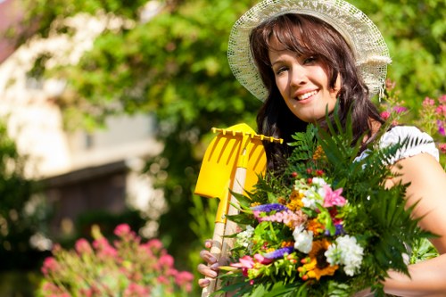 A woman standing in a well-maintained back garden during daytime, holding a yellow garden tool in one hand and a large colorful bouquet of flowers and lush green foliage in the other. The garden features a neatly trimmed lawn with dense, vibrant green grass, bordered by flower beds filled with blooming plants of various colours such as pink, white, and purple. In the background, there are tall trees and bushes providing natural privacy and shade, with sunlight filtering through the leaves. The scene captures a peaceful outdoor environment typical of a suburban area in Chiswick, with a focus on garden beauty and maintenance. The woman wears a white sun hat and a bright, cheerful expression, indicating outdoor gardening activity, and the setting reflects a cared-for garden space suitable for professional landscaping and garden services.
