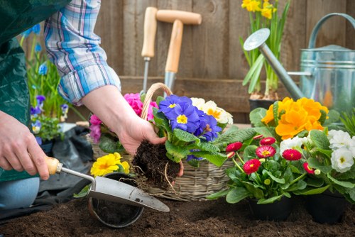 A person engaged in planting flowers in a garden bed, with visible bare soil and a variety of colourful flowering plants including primroses and violas. The individual is wearing a plaid shirt and is using a small trowel to carefully position a flowering plant with purple and yellow blooms into the soil. In the background, garden tools such as a watering can, a watering can with a yellow daffodil, and a wooden garden fence are visible, indicating a well-maintained outdoor space typical of a residential garden in Chiswick. The scene showcases a neatly arranged garden area with a mix of flower pots and garden accessories, emphasizing the importance of detailed landscaping and planting services offered by Landscapers Chiswick for enhancing outdoor environments with vibrant, natural planting layouts.