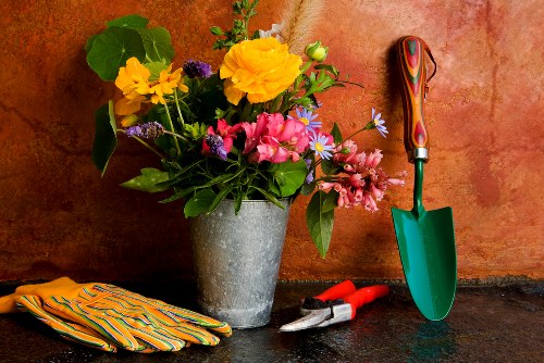 The image features a stone or concrete surface with a backdrop of a reddish-brown brick or textured wall, suggesting an outdoor or garden shed setting. In the foreground, there is a large, rustic grey flower pot filled with a mixed arrangement of vibrant flowers, including yellow, pink, and purple blooms, complemented by lush green foliage. To the right of the flower pot, a small green-handled trowel with a colorful grip is inserted into the surface, indicating gardening activity. Nearby, a pair of red-handled pruning shears and a yellow-striped gardening glove are placed on the surface, suggesting recent planting or maintenance work. The scene is well-lit with natural daylight, evoking an early morning or late afternoon outdoor environment. The combination of tools, gloves, and flowering display reflects typical garden care activities, aligning with services offered by landscapers in Chiswick and illustrating the process of flower bed creation or outdoor garden maintenance within a typical UK garden setting.