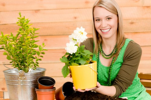 The image depicts a woman with shoulder-length blonde hair smiling while holding a yellow flowerpot containing a flowering plant with white blossoms and green foliage. She is seated outdoors on a patio or garden area with a wooden fence or wall in the background. In front of her, there are various gardening items including a metal bucket with a small, leafy green shrub, an empty black plant pot, and an orange terracotta pot. The setting suggests a well-maintained outdoor space suitable for gardening activities, with natural lighting and a casual, tranquil atmosphere. The scene highlights gardening tasks such as pot planting and plant maintenance, aligning with professional gardening services in areas like Chiswick and the broader London region, emphasizing outdoor gardening and landscape care.