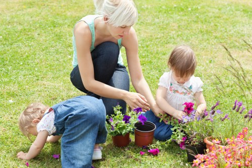 A woman with short blonde hair, wearing a light blue sleeveless top and dark jeans, is kneeling on a well-maintained, lush green lawn, engaging with two young children in a garden setting. One child, dressed in white and blue, is leaning forward on hands and knees, exploring the grass and flower petals, while the other, wearing a white t-shirt with a floral pattern and blue jeans, is sitting nearby, gently tending to a potted plant with vibrant purple flowers. Surrounding them are various flower pots with colourful blooms, some placed directly on the grass, adding natural tones of purple, pink, and green to the scene. The garden area is spacious with a foreground of evenly cut grass, and background elements include additional potted plants, suggesting a well-kept outdoor space suitable for family gardening activities. The natural daylight indicates a clear day, emphasizing the vivid colours of the flowers and greenery, aligning with gardening and outdoor maintenance services offered by Landscapers Chiswick in the local Chiswick area, near the W4 postcode.