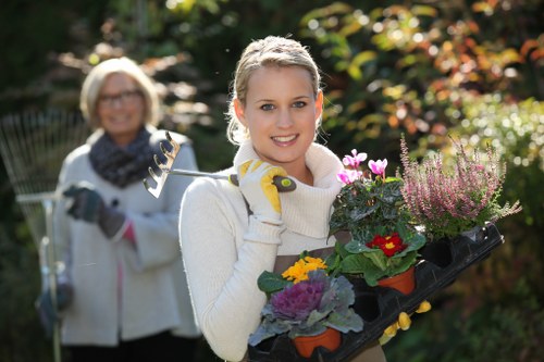 A young woman with blonde hair, wearing a white sweater and gardening gloves, is holding a black tray filled with colorful flowering plants, including red, purple, and yellow blooms, in a lush garden. The garden features a well-maintained green lawn in the foreground, bordered by flower beds and shrubbery with dense foliage. In the background, an older woman wearing glasses, a white jacket, and a patterned scarf is smiling and holding a garden tool, with a backdrop of trees and bushes displaying a mix of green and autumnal colours. The scene suggests outdoor gardening activity on a bright, sunny day with natural light highlighting the vibrant plant colours and the healthy, textured grass. The setting appears to be a well-kept private or community garden in Chiswick, with natural surfaces of soil, paving, and flourishing greenery, aligning with professional gardening and landscaping services offered by Landscapers Chiswick.